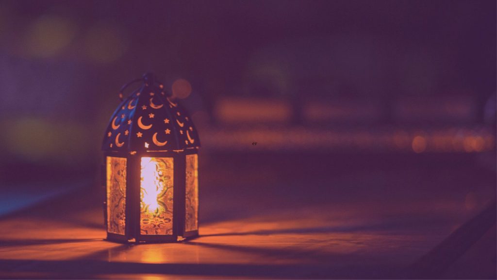 A decorative lantern with star and moon cutouts emits a warm glow on a reflective surface in a dimly lit setting.