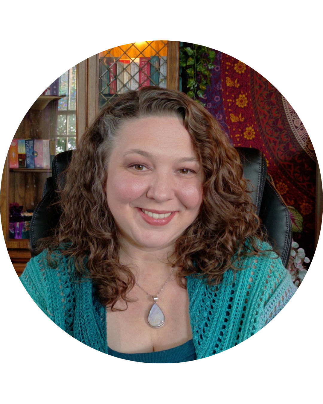 A woman with curly brown hair sits in a chair, smiling at the camera. She wears a turquoise sweater and a teardrop-shaped pendant. Books and colorful décor are in the background.